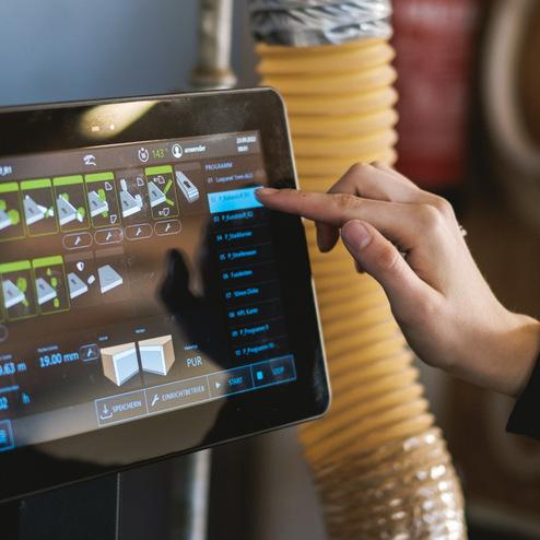 Nahaufnahme einer Hand, die einen Touchscreen bedient. Der Bildschirm zeigt Programme und Einstellungen einer Produktionsmaschine mit technischer Benutzeroberfläche.