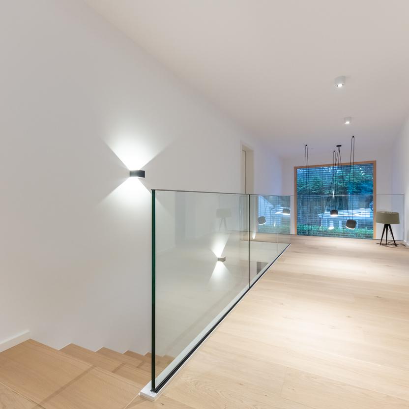 Heller, minimalistischer Flur mit Holzfußboden, Glasgeländer, Wandbeleuchtung und Blick auf große Fenster mit Hängelampen.