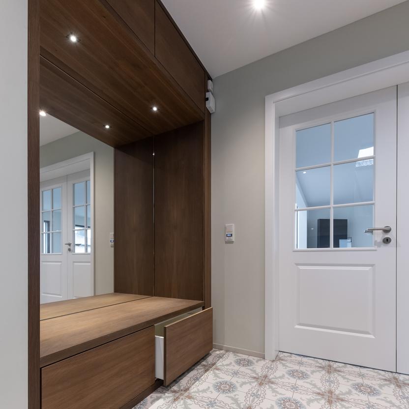 Stilvolle Garderobe nach Maß in einem Eingangsbereich Maßgefertigte Garderobe in Holzoptik mit Schubladen und einem Sitzbereich, auf dekorativen Fliesen, neben einer weißen Tür.