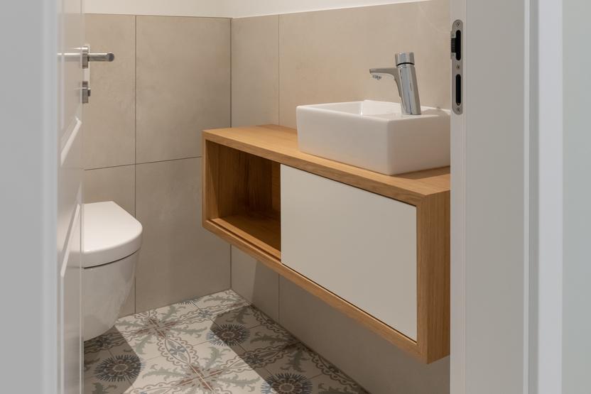 Gäste-WC mit hängendem Holzwaschtisch, modernem Waschbecken, neutralen Wandfliesen und dekorativen Bodenfliesen im floralen Muster.