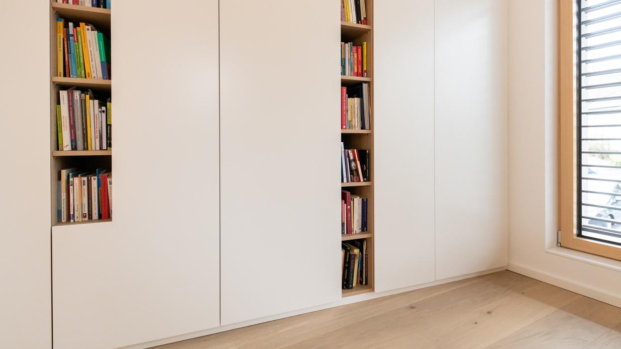 Heller, maßgeschneiderter Schrank neben einem Fenster, mit einem offenen Bücherregal voller bunter Bücher.