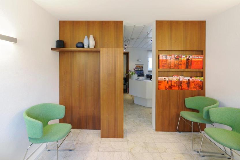 Zwei grüne Stühle im Vordergrund, mit einem Holzregal und Zeitschriften an der Wand, blickend auf eine offene Tür zum Empfangsbereich.