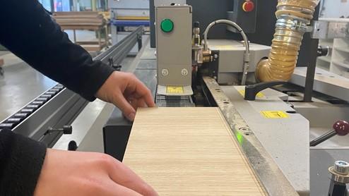 Bearbeitung von Holz an einer Kantenanleimmaschine Hand führt ein Holzstück durch eine Kantenanleimmaschine in einer Werkstatt. Die Maschine ist mit Bedienelementen und einem Display ausgestattet.