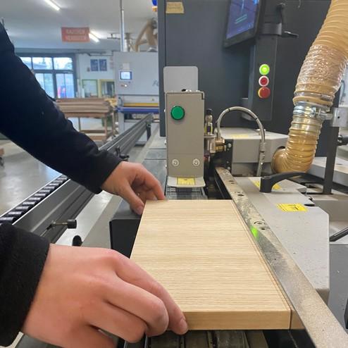 Hand führt ein Holzstück durch eine Kantenanleimmaschine in einer Werkstatt. Die Maschine ist mit Bedienelementen und einem Display ausgestattet.